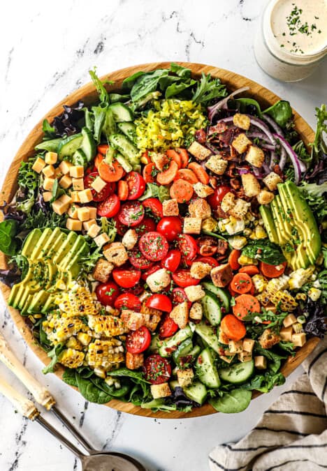 showing how to make garden salad recipe by adding tomatoes, cucumbers, carrots, red onions, avocados, croutons and cheese to a bowl