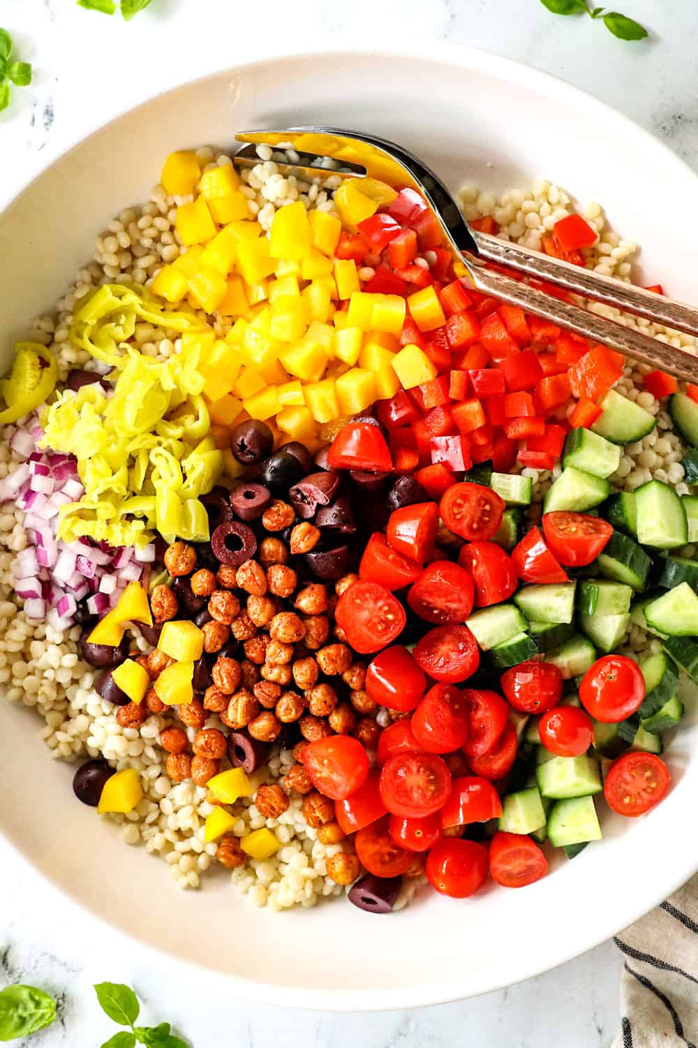 top view showing how to make couscous salad recipe by adding pearl couscous, tomatoes, cucumbers, chickpeas, bell peppers, red onions and olives to a bowl and tossing to combine