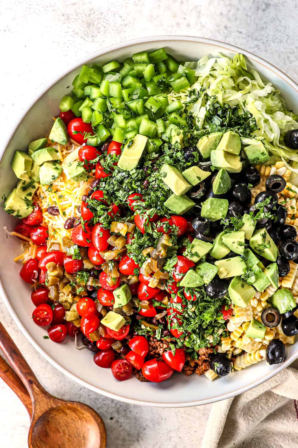 showing how to make taco pasta salad recipe by adding pasta, ground beef, tomatoes, corn, black beans, bell peppers, avocados, and olives to a bowl