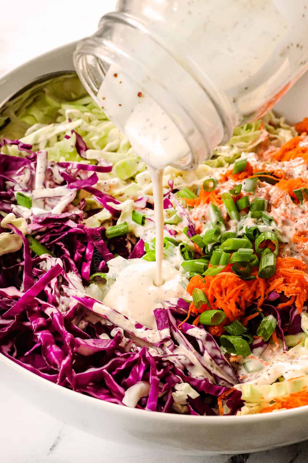 showing how to make coleslaw recipe by pouring coleslaw dressing over the cabbage, carrots and green onions
