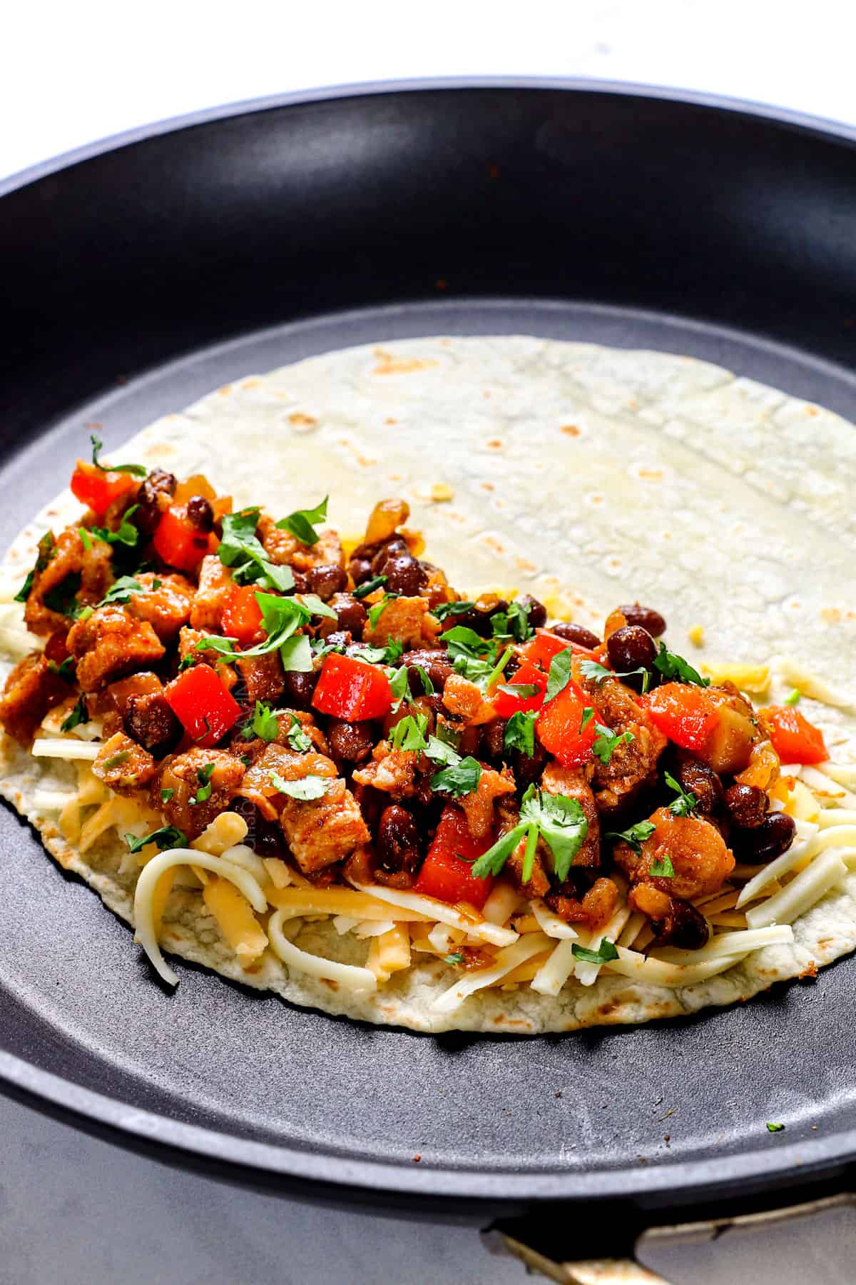 showing how to make chicken quesadilla by adding the tortilla to a skillet and topping half of it with cheese, chicken, veggies and cilantro