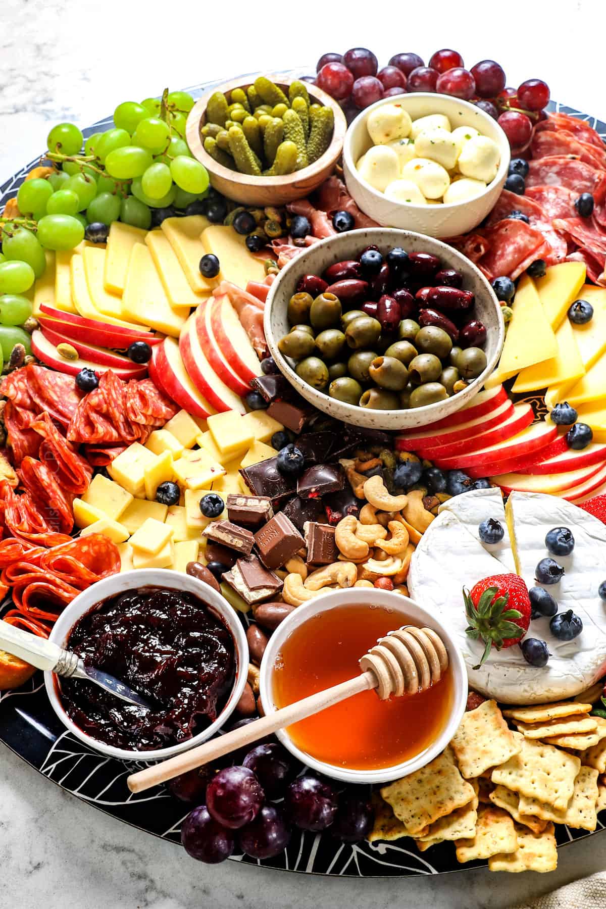 a meat and cheese charcuterie board with salami, prosciutto, Havarti, Gouda, brie, goat cheese, grapes, strawberries, blueberries, apples, honey, jam and nuts to a board