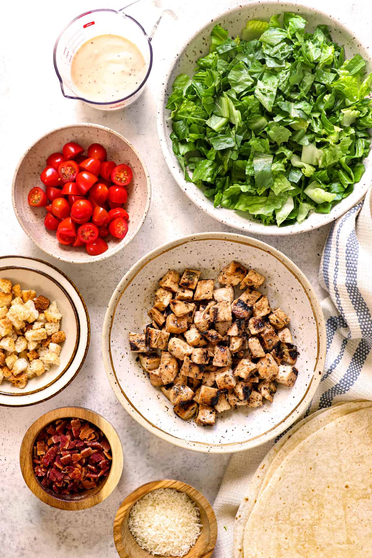 showing ingredients for chicken Caesar Wrap lined on the table: grilled chicken, Romaine lettuce, tomatoes, croutons, bacon, Parmesan and Caesar dressing