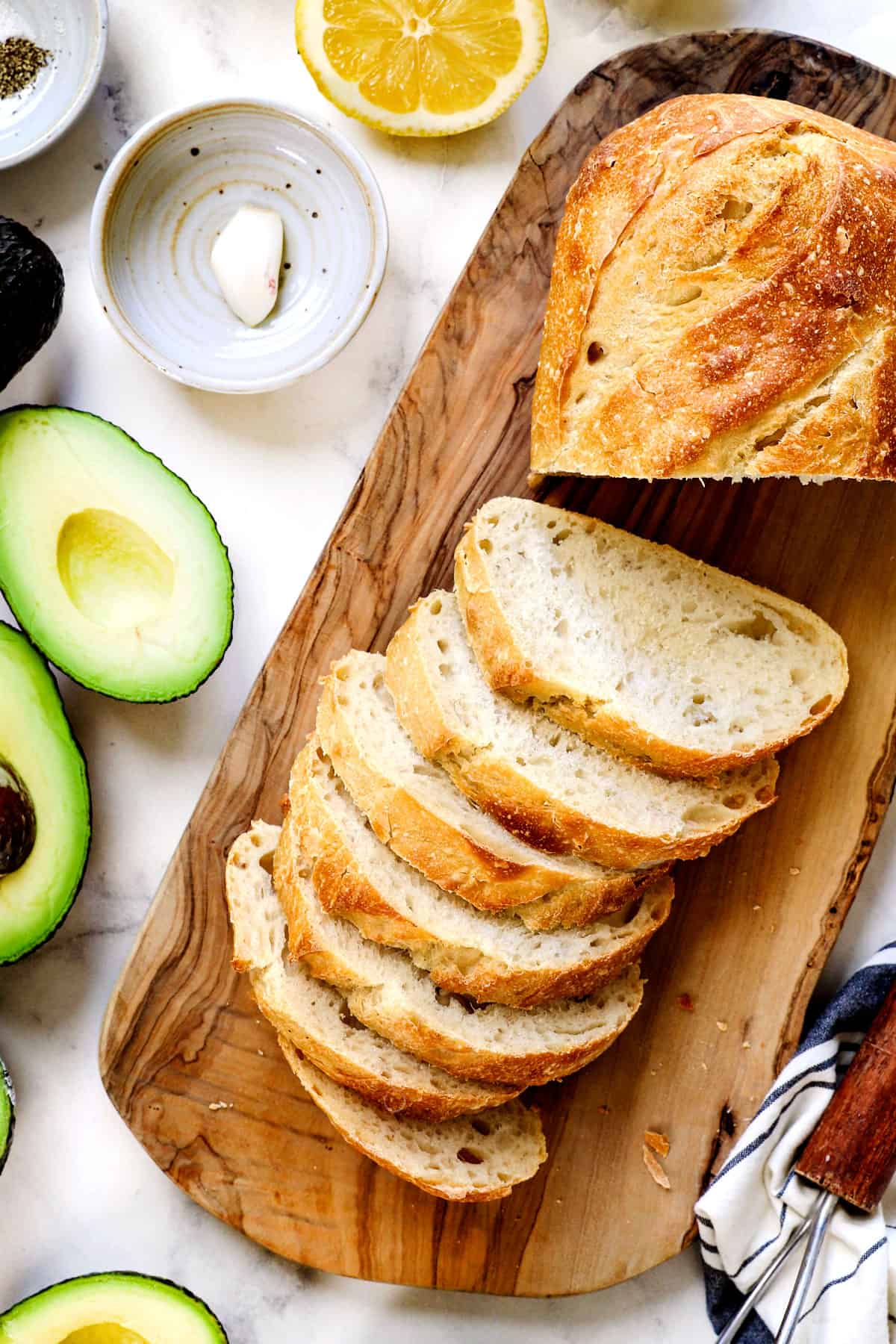 top view showing the ingredients for avocado toast: sourdough bread on a cutting board, avocados, lemon juice, garlic, salt and pepper