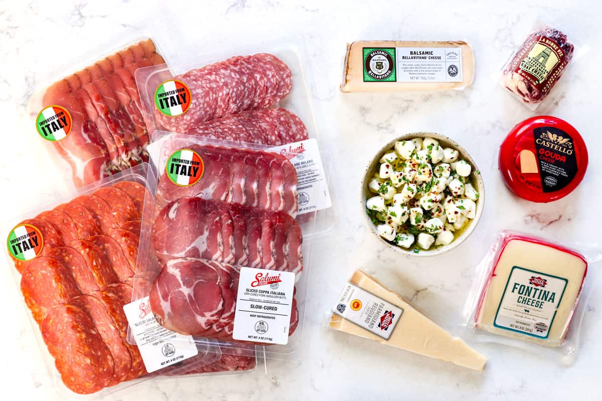 top view showing meats and cheeses for antipasto platter: โข Parmigiano Reggiano, Provolone, Fontina, Balsamic BellaVitano, marinated mozzarella balls, cheese log, Genoa salami, Soppressata, prosciutto, dried coppa