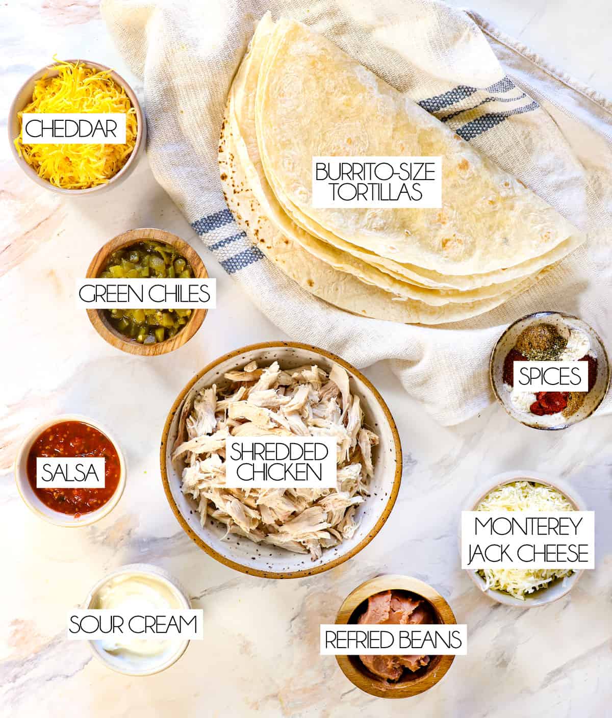 top view of chimichanga recipe: burrito size tortillas, cheddar cheese, Monterey Jack cheese, shredded chicken, green chiles, salsa, sour cream, refried beans, spices