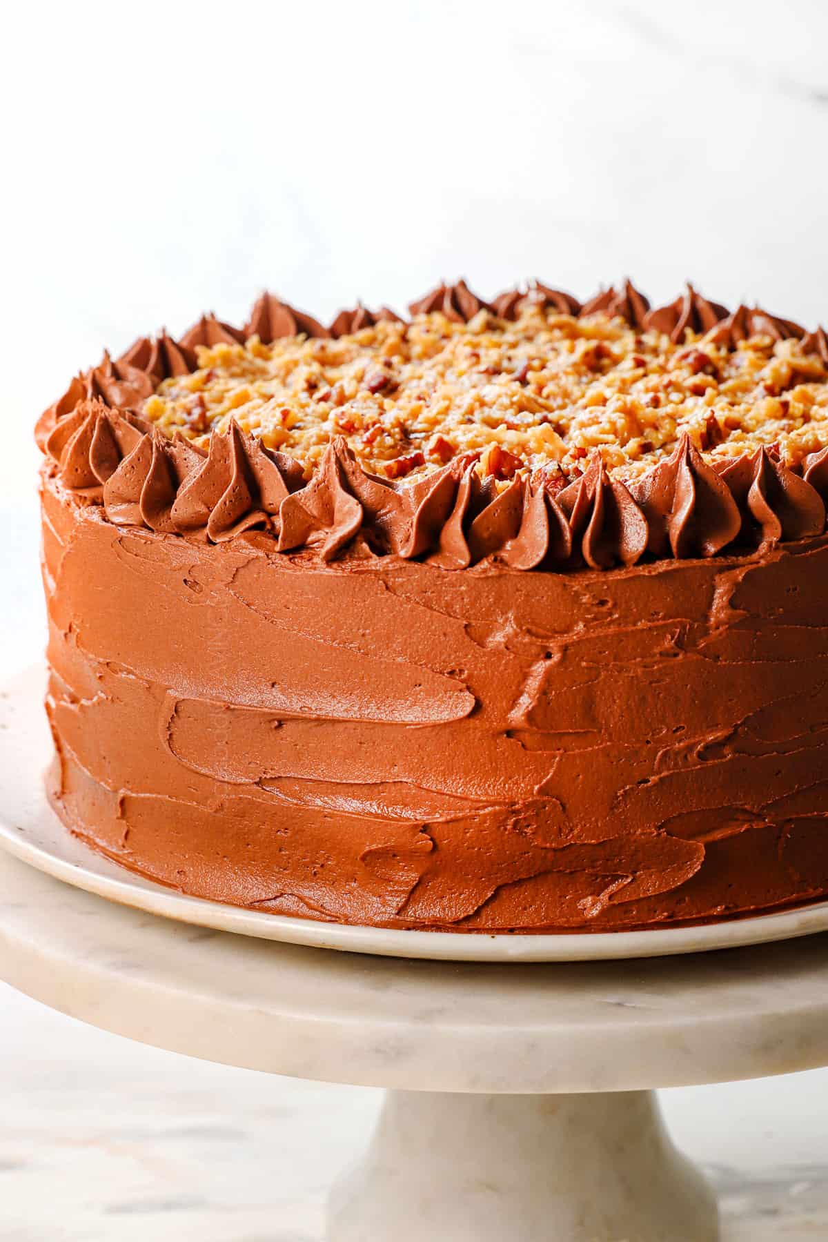 German Chocolate Cake on a pedestal showing that it's frosted all over with buttercream frosting, with a top layer of Chocolate German Frosting