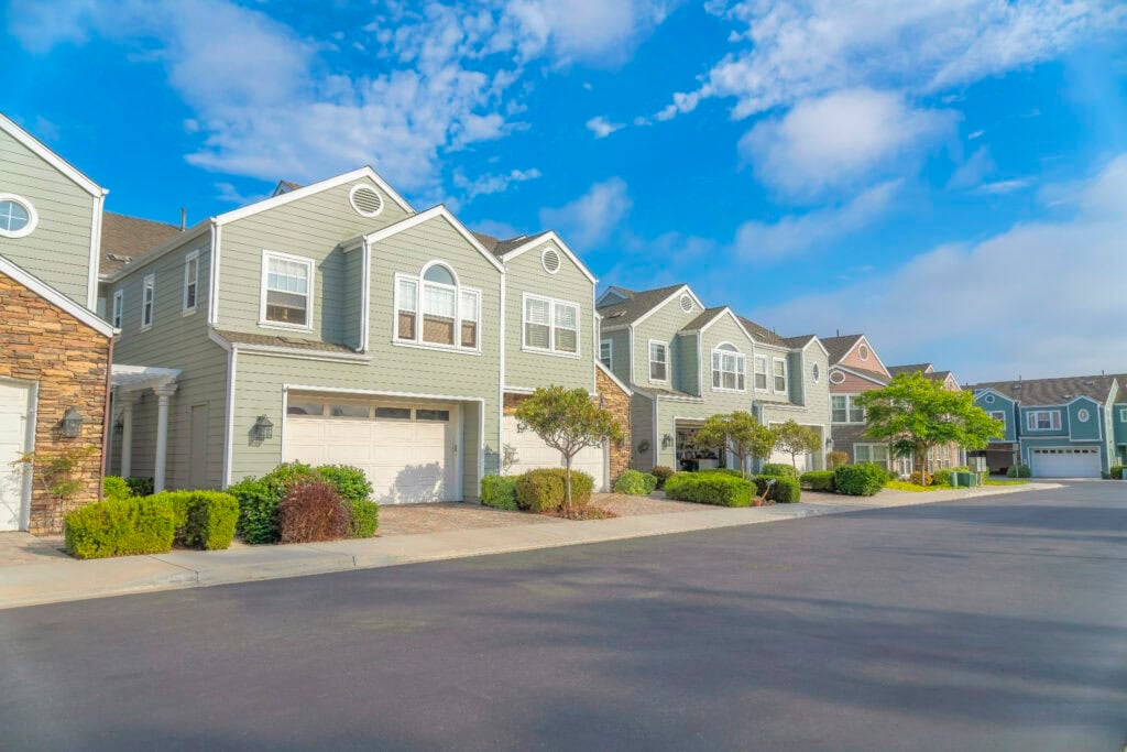 Duplex houses with green wood vinyl sidings at Carlsbad, San Diego, California. There are white garage doors with concrete driveway near the plants at the front. how to make $30,000 a month with real estate