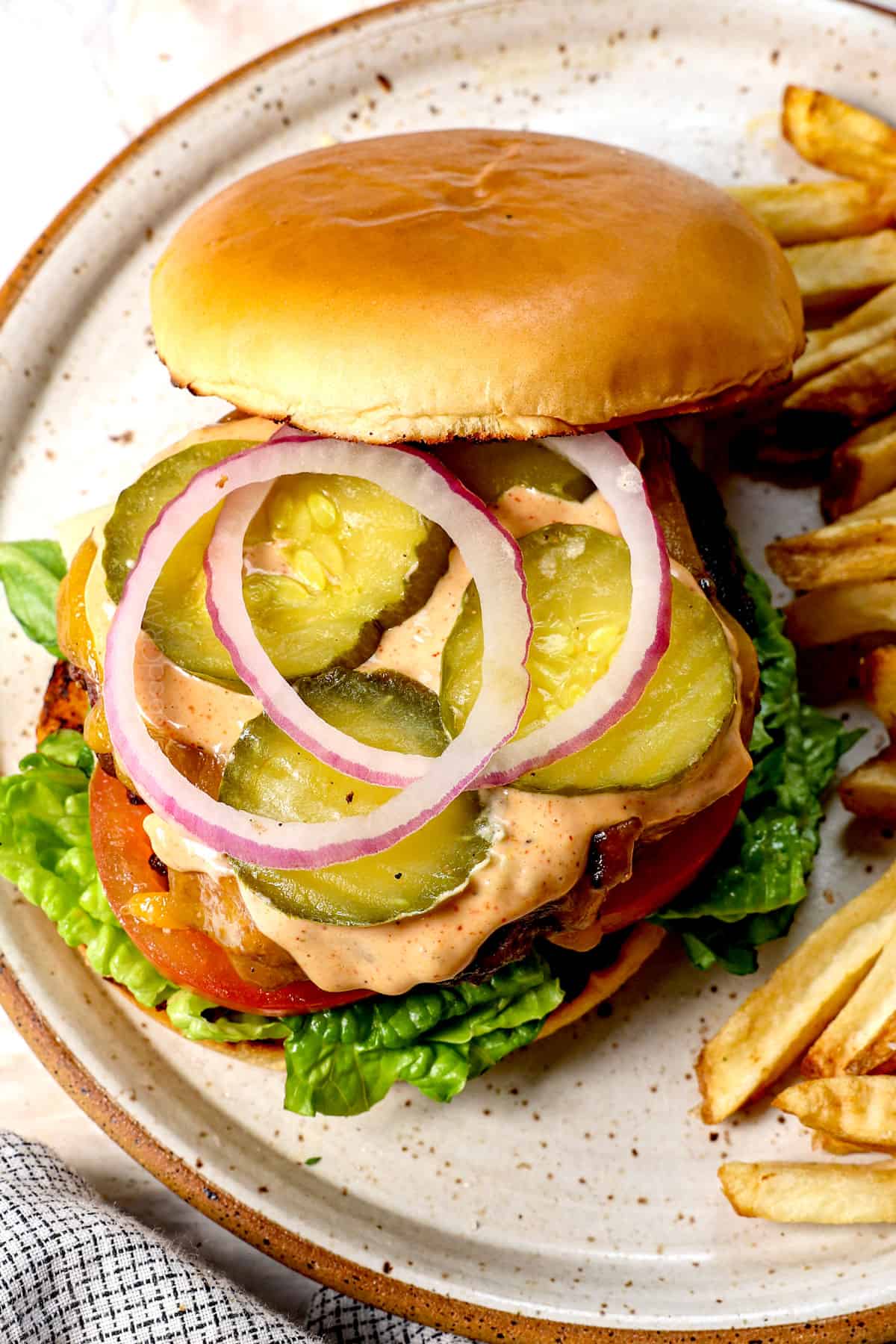 showing how to make a cheeseburger by assembling with the buns, lettuce, tomatoes, burger, pickles, red onions, then topping with the top bun