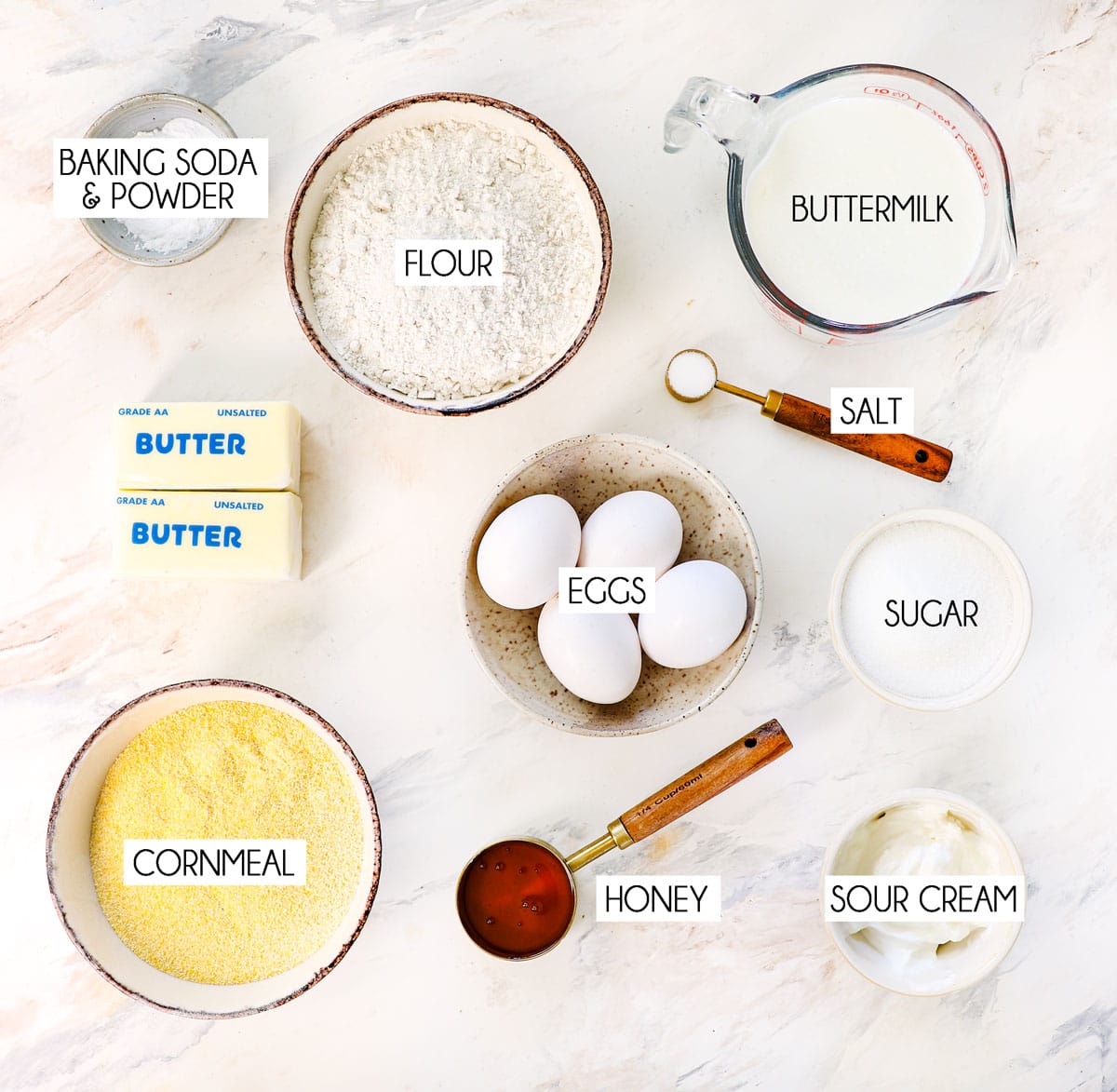 top view of cornbread ingredients: cornmeal, flour, sugar, eggs, buttermilk, butter, baking soda, baking powder, salt, sour cream and honey