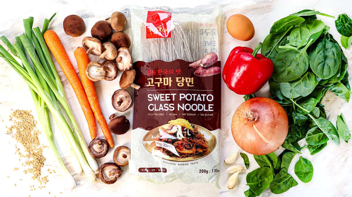top view of japchae ingredients for the stir fry portion showing what sweet potato noodles look like, along with yellow onion, green onions, carrots, red bell pepper, spinach, garlic, and shiitake mushrooms