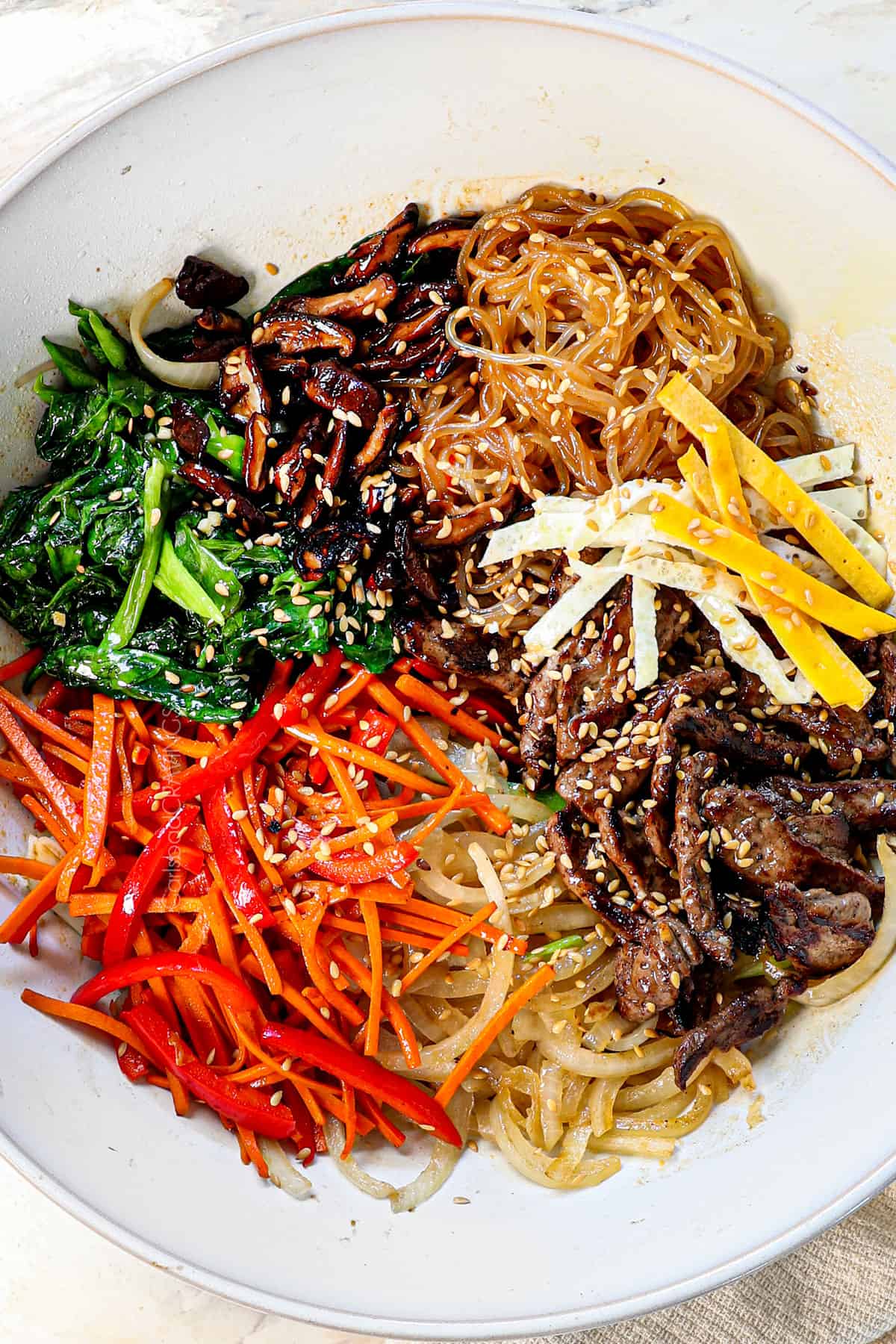 top view of Japchae ingredients in a bowl after being cooked: glass noodles, beef strips, red bell pepper, carrots, onions, shiitake mushrooms and spinach