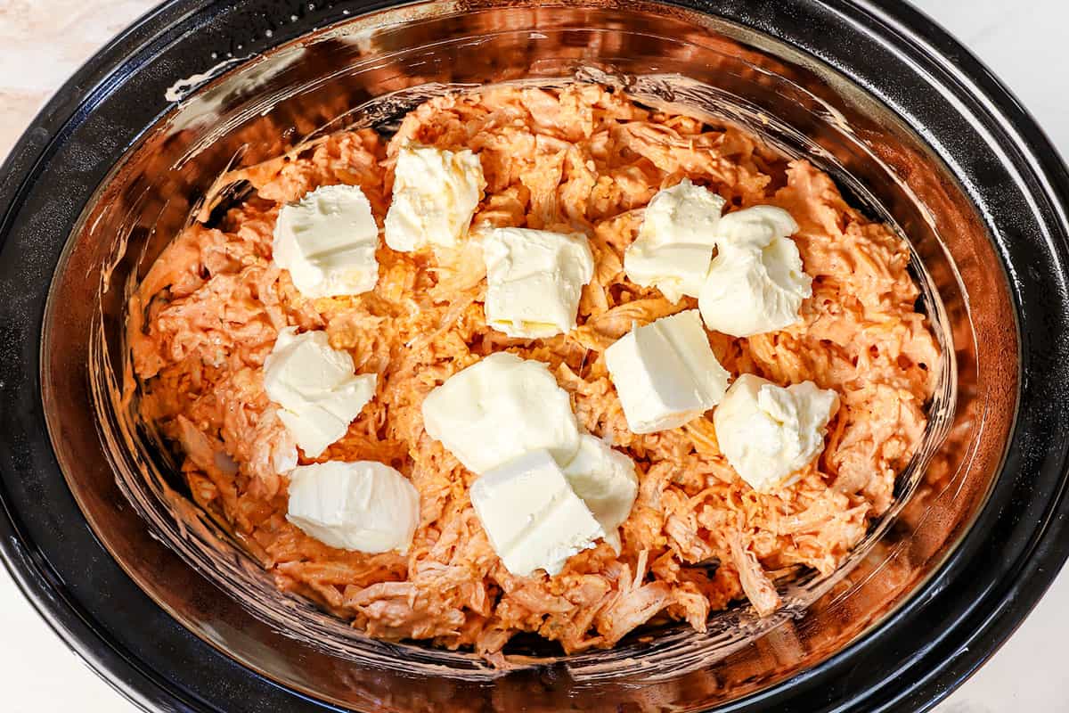 showing how to make crockpot buffalo chicken dip by adding cubed cream cheese on top of the chicken mixture in the crockpot